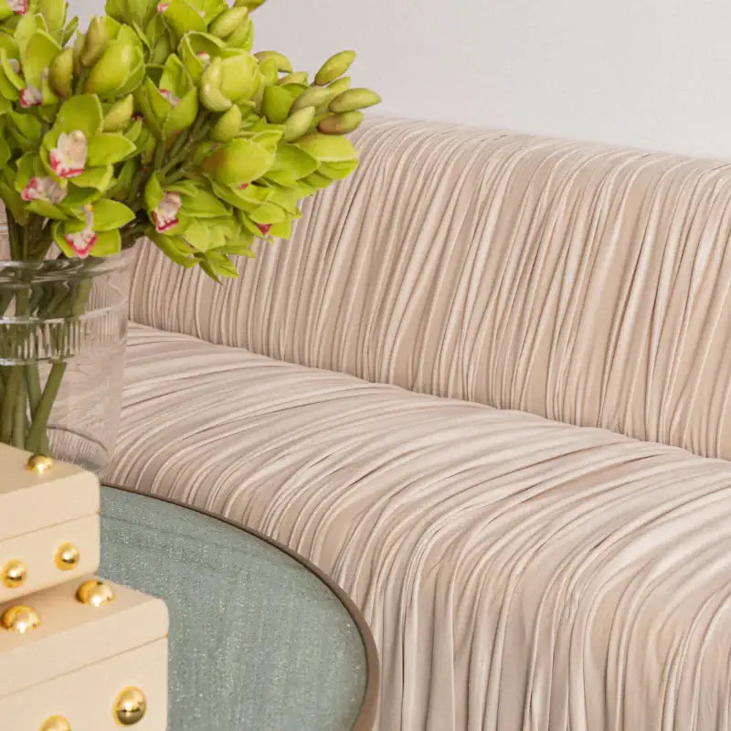 A close-up of a beige upholstered sofa with vertical pleats, styled with a vase of green orchids and a round mint-green side table in a bright interior setting.