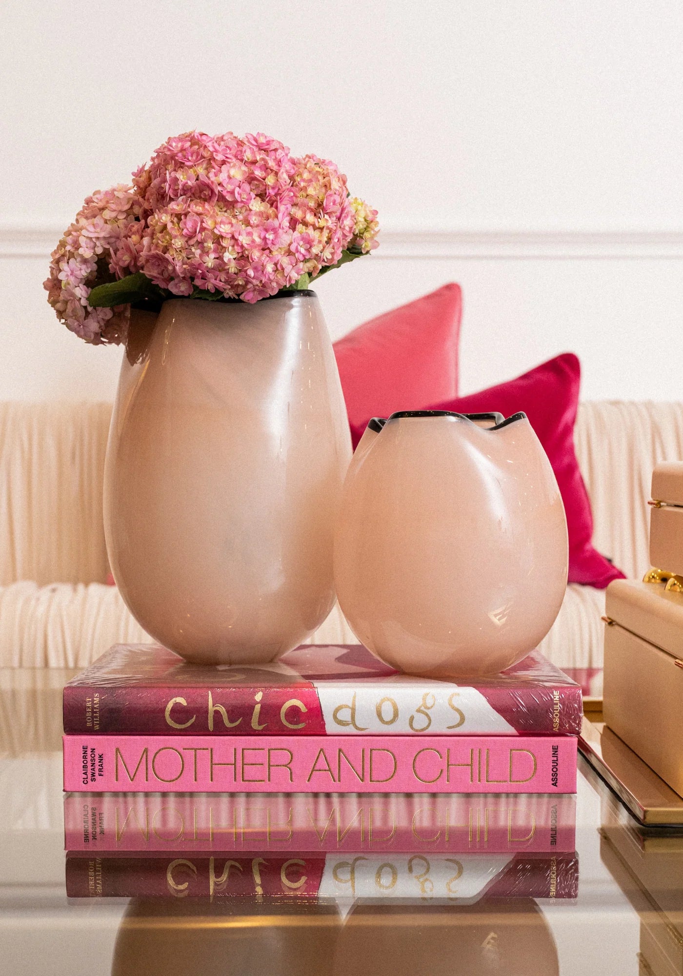 Two matte blush pink ceramic vases, one tall and one short, styled on top of a pink and white book with pink hydrangea flowers in the taller vase, set against a cream sofa with pink pillows.