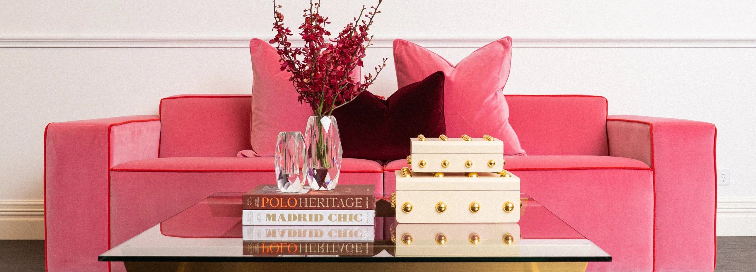 A bright pink velvet sofa in a styled room setting, decorated with pink and dark red pillows, a glass vase with red branches, a stack of books, and a decorative box on a glass coffee table.