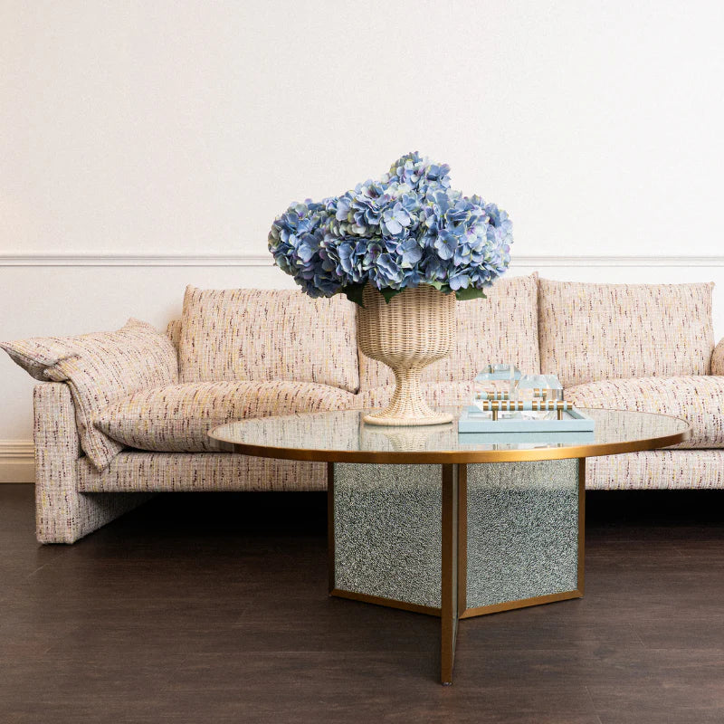 A round coffee table with a glass top, gold-tone frame, and textured silver-and-gold base, styled in a living room with a beige patterned sofa and a large vase of blue hydrangeas on top.