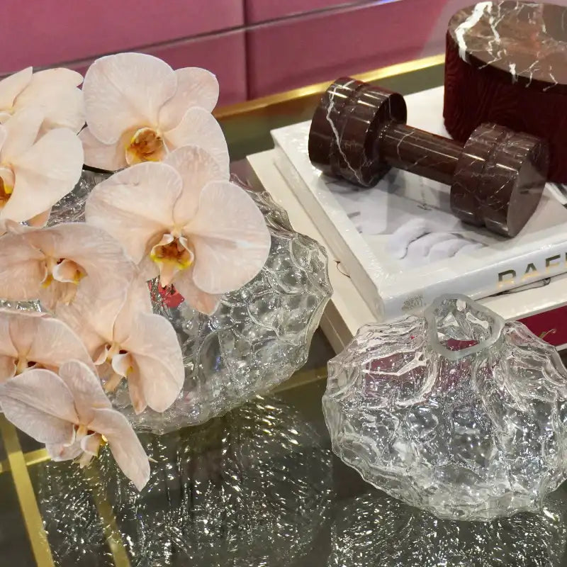 A styled tabletop scene with pale pink orchid flowers in a textured clear glass vase, a dark brown marble dumbbell resting on stacked white books, and another textured clear glass object in the foreground.