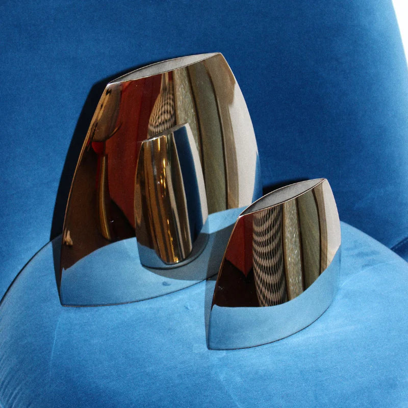 Two modern abstract metallic wall art pieces with reflective chrome surfaces, displayed on a blue velvet surface.