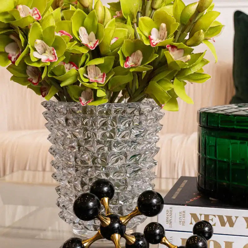 Green artificial orchid flowers with white and pink centers arranged in a textured clear glass vase, set on a table with books and a green container in a styled indoor setting.