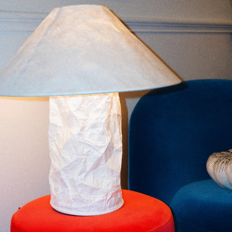 A table lamp with a crumpled white paper base and a light gray fabric shade, sitting on a red cylindrical side table next to a blue armchair.