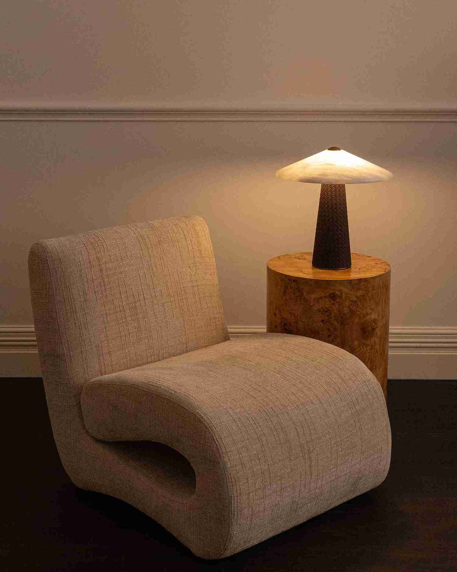 A modern low-profile armchair in textured beige fabric, positioned next to a rustic wooden side table holding a glowing mushroom-shaped table lamp against a neutral wall.