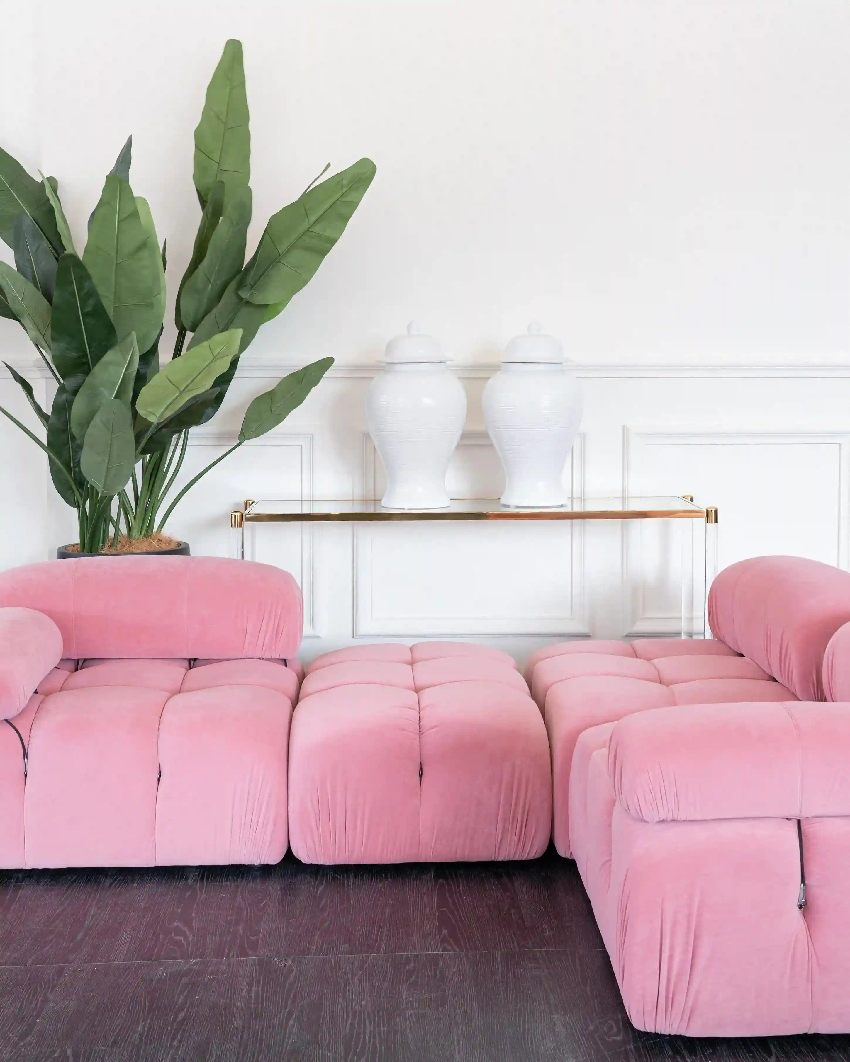 A pink tufted modular sofa set in a bright living room, with a matching ottoman in the center and a large green potted plant and white ceramic vases on a glass shelf in the background.
