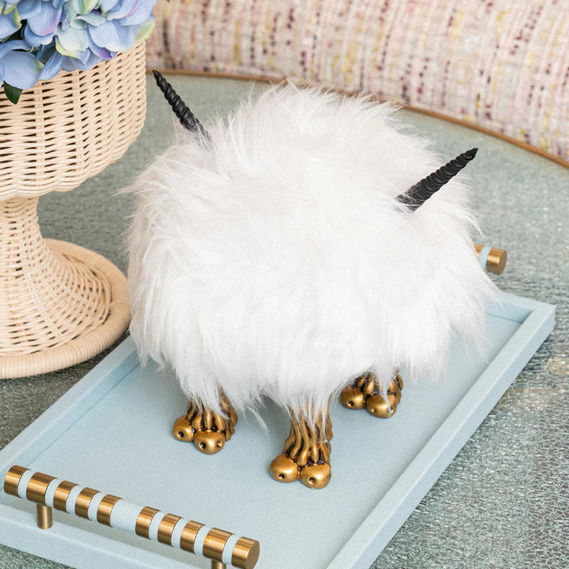 A fluffy white decorative unicorn figurine with black horns and gold hooves, displayed on a light blue tray in a styled interior setting.