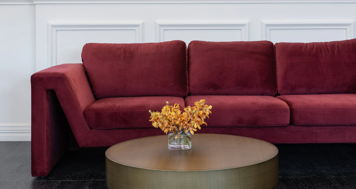 A modern three-seater sofa upholstered in deep burgundy velvet, positioned behind a round metallic gold coffee table holding a vase of yellow flowers.