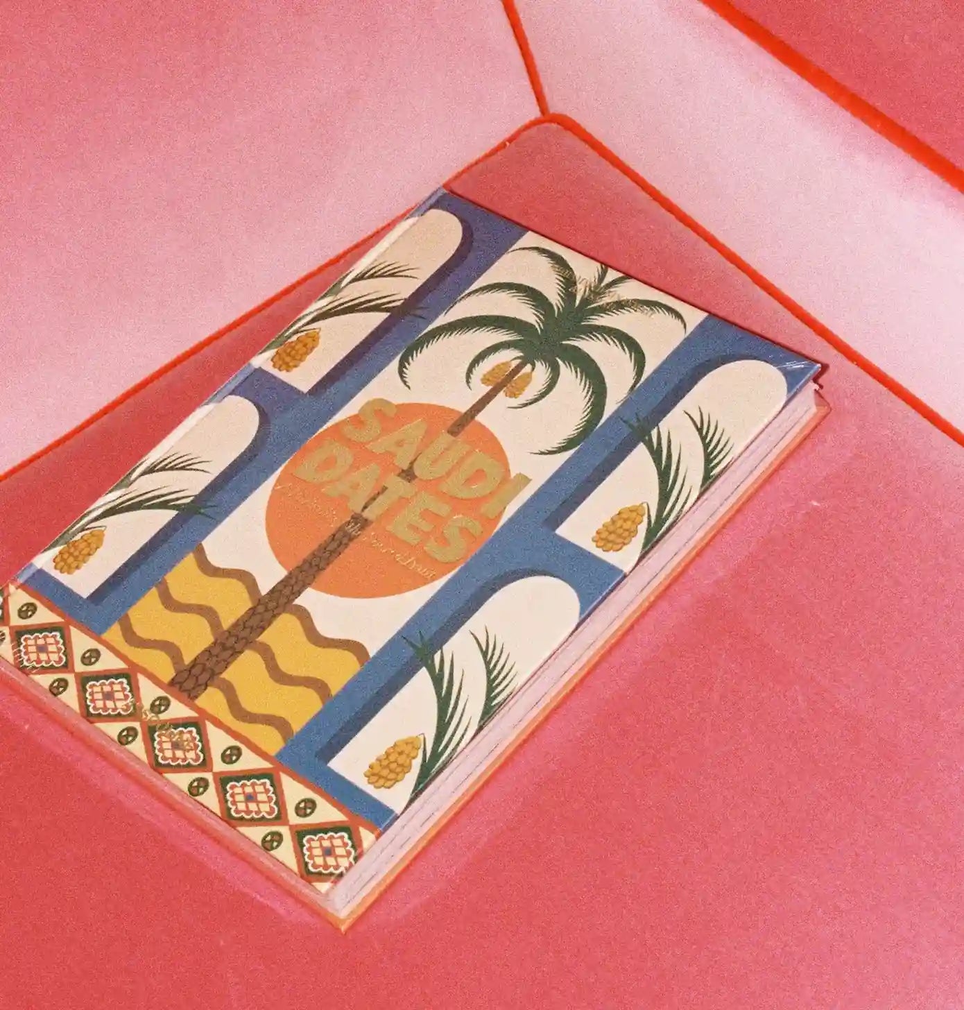 A notebook titled 'SAUDI DATES' with a colorful retro pattern of palm trees, suns, and geometric borders, resting inside a pink envelope.