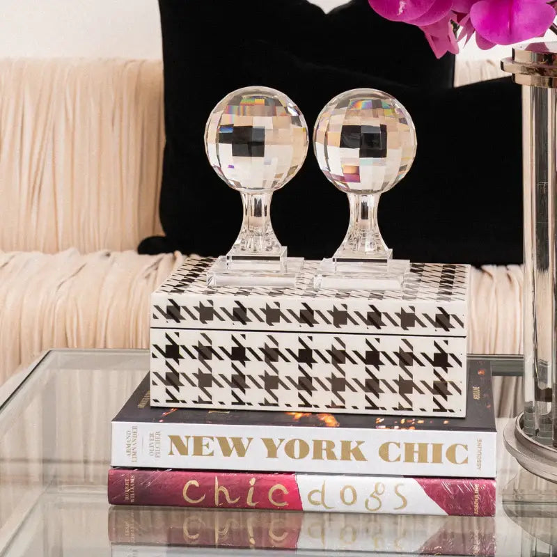 A decorative rectangular box with a black-and-white houndstooth pattern sits on stacked books on a glass table, with two clear faceted crystal globes on top. A black pillow, cream pleated upholstery, and a pink orchid are visible in the background.