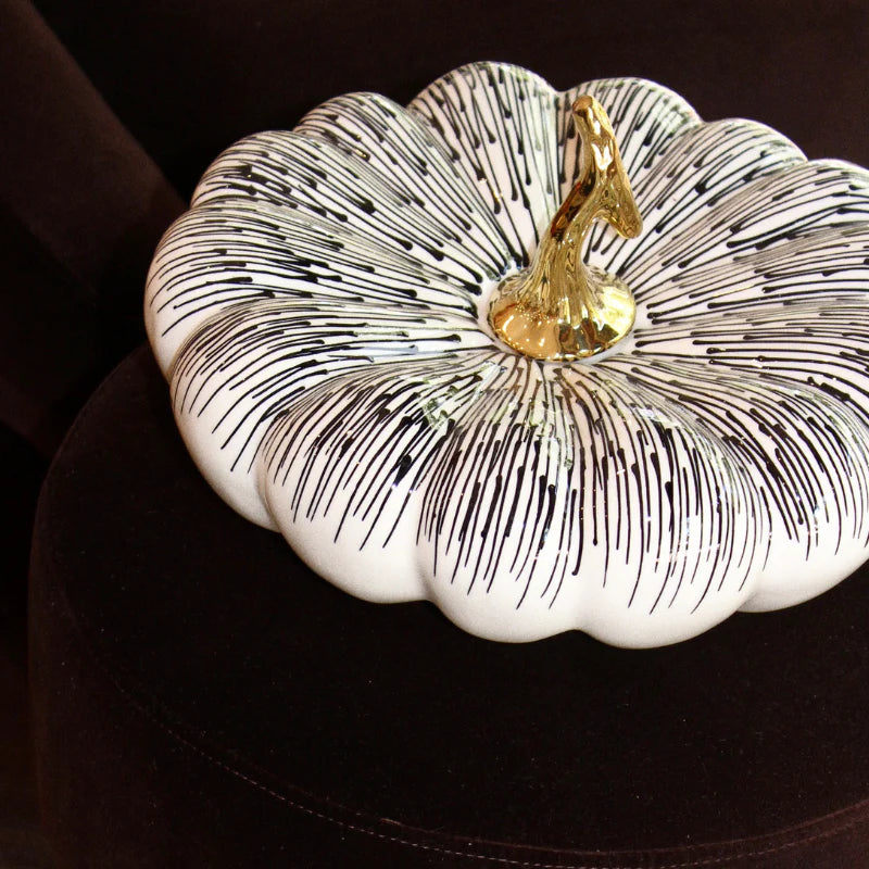 A decorative pumpkin-shaped container or box with a white base, black linear stripe pattern across the surface, and a metallic gold top handle, shown on a dark background.