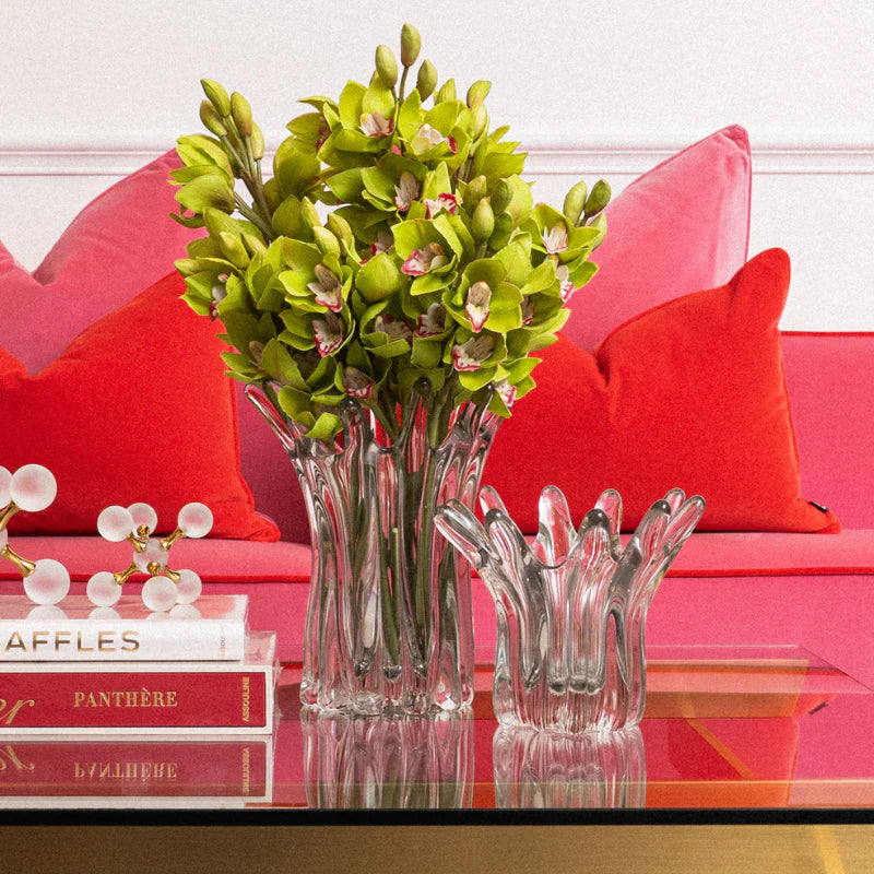 A green orchid floral arrangement in a tall ribbed clear glass vase, displayed on a glass-topped table in front of a bright pink sofa with red pillows. A matching clear glass vase with sculptural leaf-like stems sits beside it.