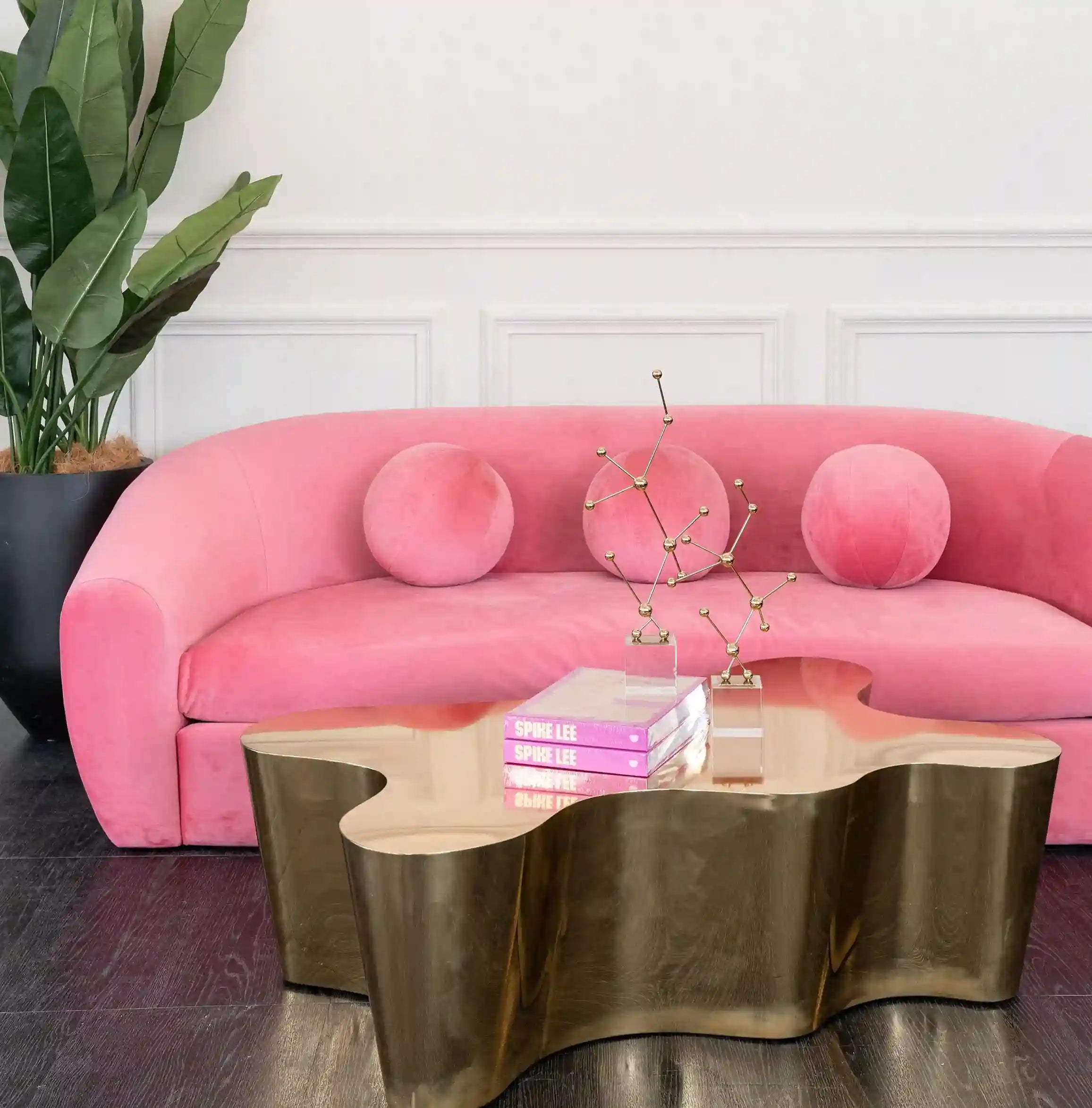 A pink velvet sofa with three round pink pillows in a styled living room setting, featuring a metallic gold wavy coffee table and a potted plant against a white paneled wall.