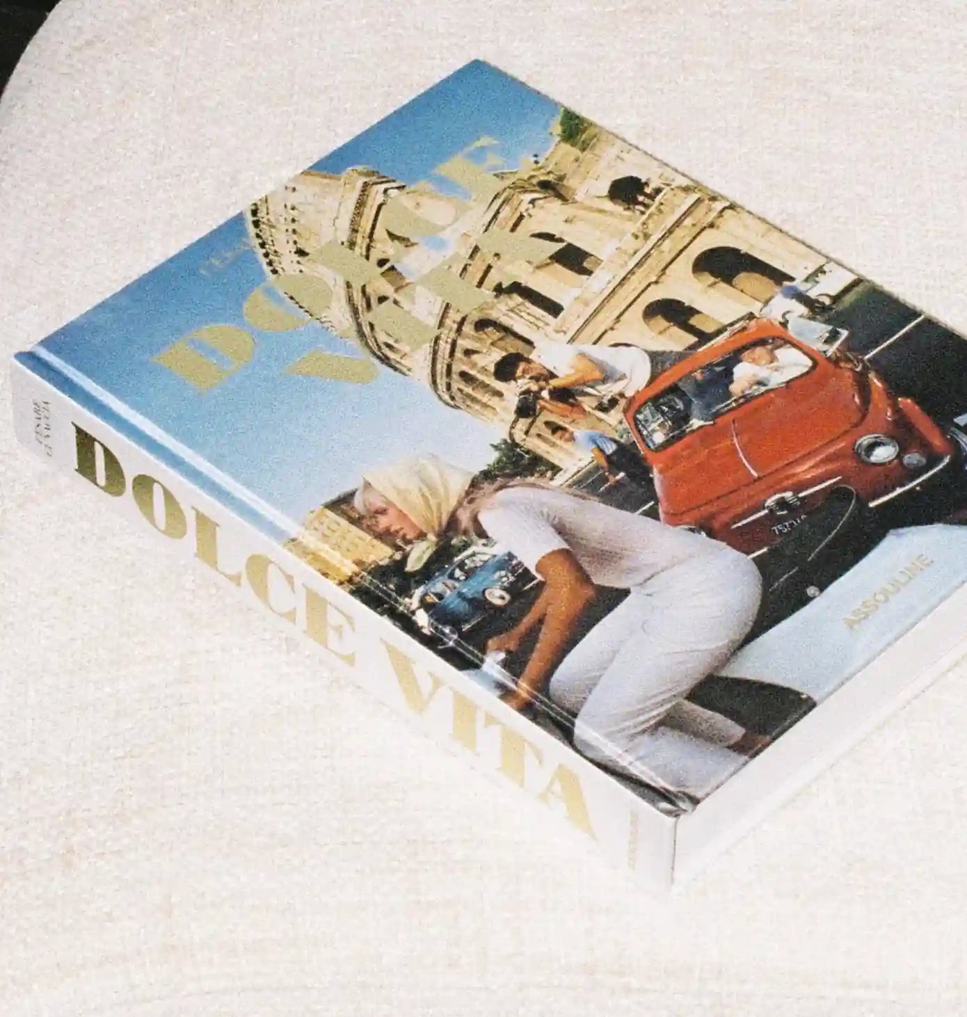 A hardcover book titled 'Dolce Vita' by Anna Sui lies on a textured off-white fabric surface. The cover features a vibrant photograph of a woman in white clothing leaning over a classic red convertible car, with the Colosseum in Rome in the background under a blue sky.