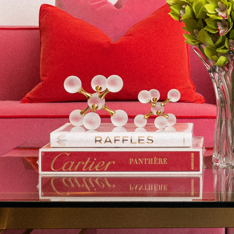 A decorative crystal and gold geometric sculpture set displayed on top of stacked books on a glass table, with a pink sofa, red pillow, and green flowers in the background.