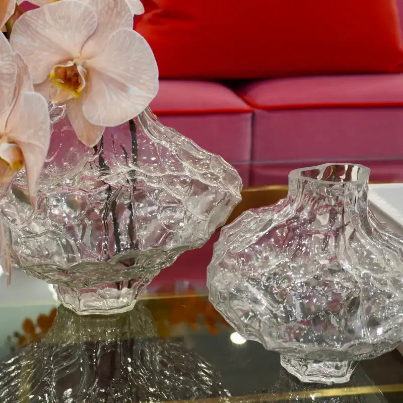 Two clear textured glass vases on a reflective tabletop, with pale pink orchids in the larger vase and a red upholstered sofa in the background.