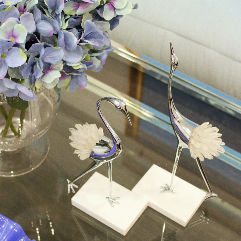 Two decorative silver crane figurines with white feathered details and white square bases, displayed on a glass tabletop beside a vase of blue and purple flowers.