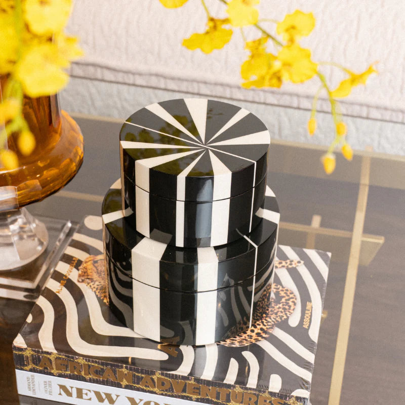 Three stacked round decorative boxes with a glossy black and white geometric striped pattern, styled on top of books on a glass table with yellow flowers in the background.