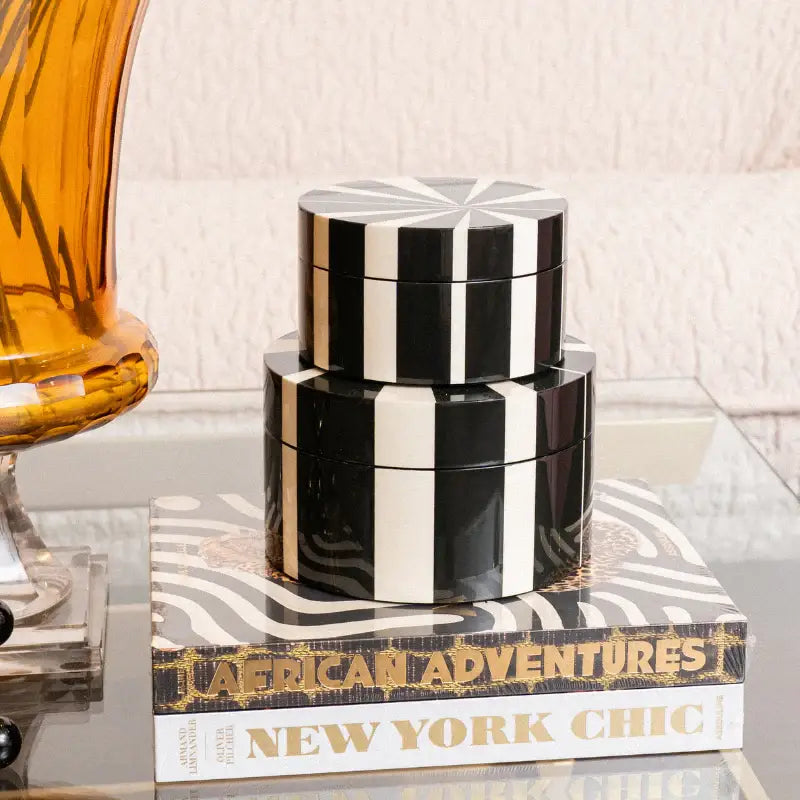 Three stacked round black and white striped cosmetic boxes on a glass table, styled on top of a book next to an amber glass vase.