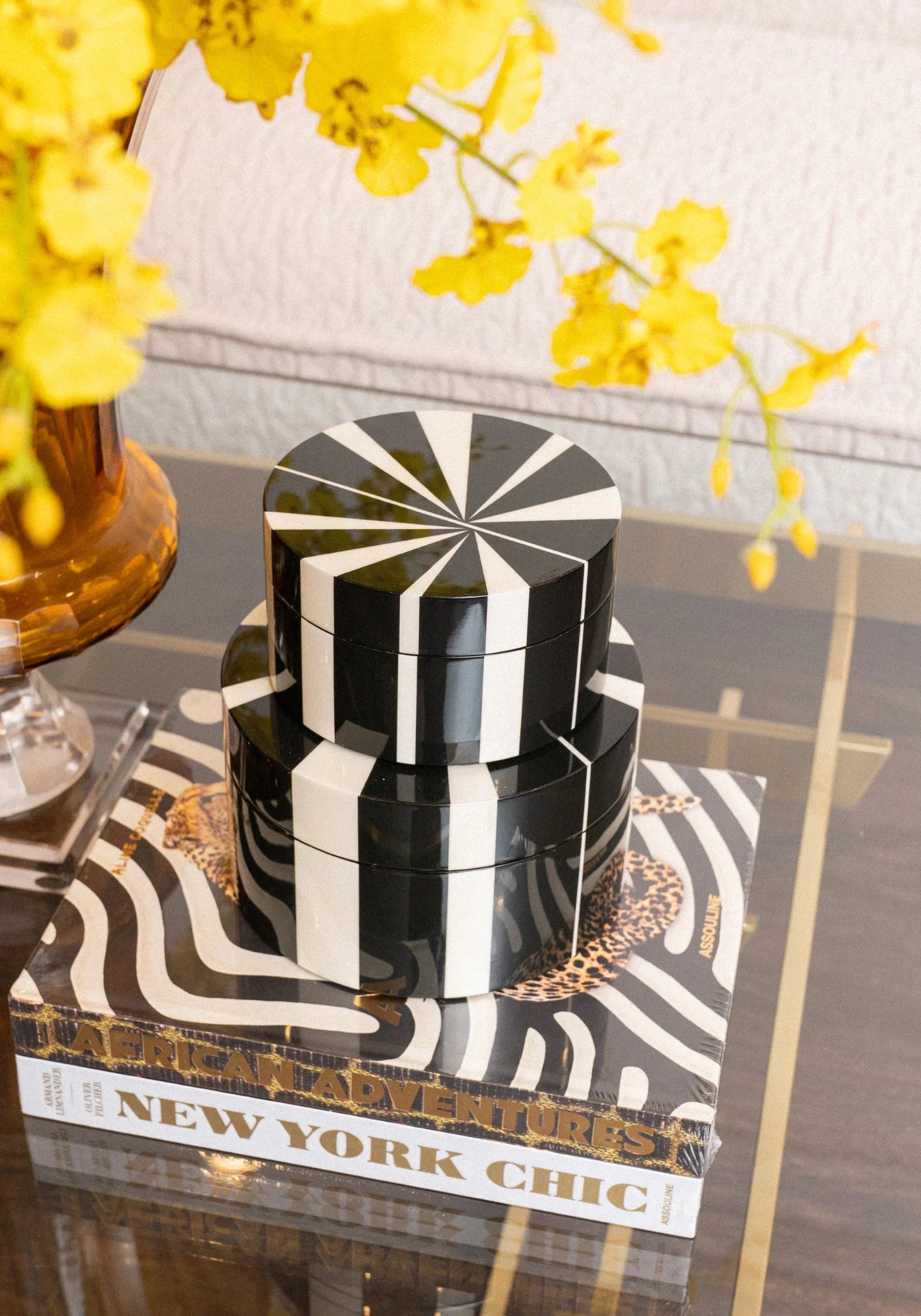 A tiered black and white striped lacquer box set styled on a zebra-print book next to yellow orchids on a glass table.