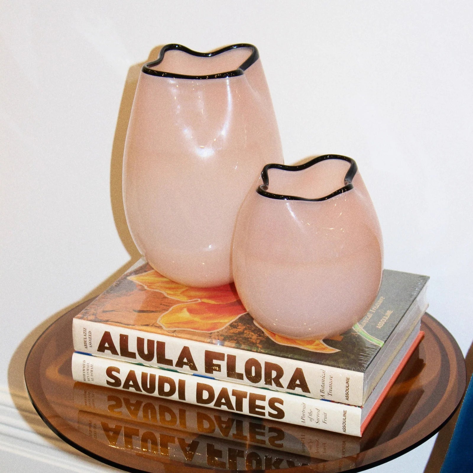 Two pale blush pink glass vases with black-rimmed scalloped openings, displayed on top of stacked books on a round reflective table against a plain light background.