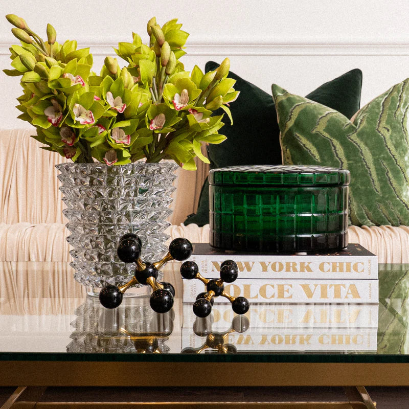 A styled interior scene showing a clear textured glass vase filled with bright yellow-green orchids on a glass table, next to a dark green ribbed glass container and books, with decorative black-and-gold orb sculpture and green pillows on a sofa in the background.