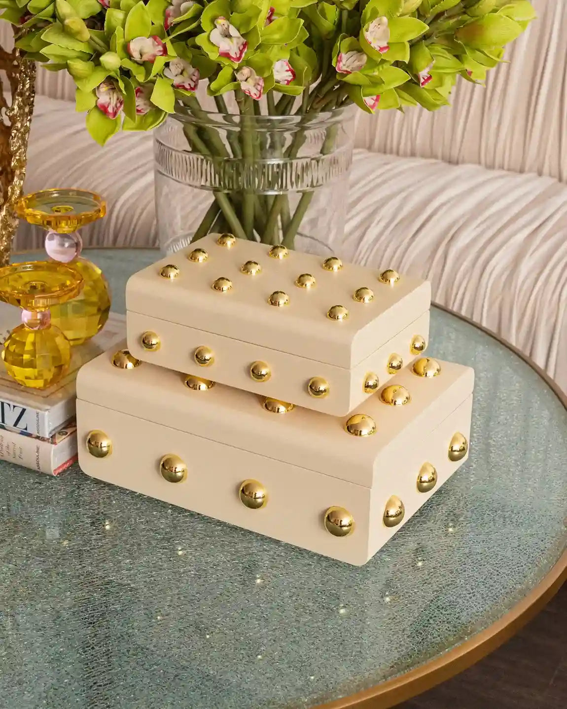 Two stacked cream-colored faux leather jewelry boxes with gold studs, styled on a round glass table with a vase of green orchids in the background.