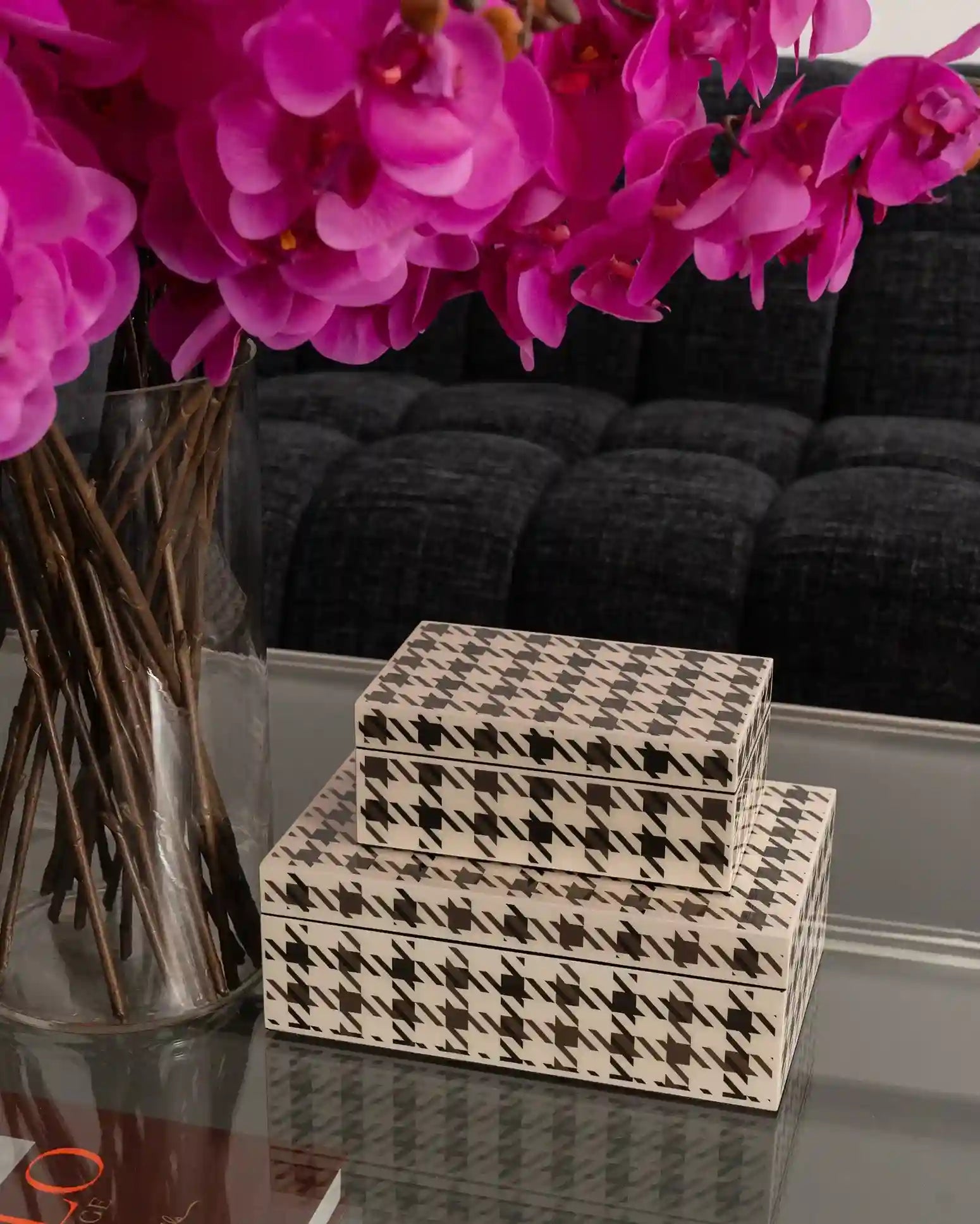 Three stacked decorative boxes with a black and white houndstooth pattern, styled on a glass table next to a vase of bright pink orchids in front of a dark gray sofa.