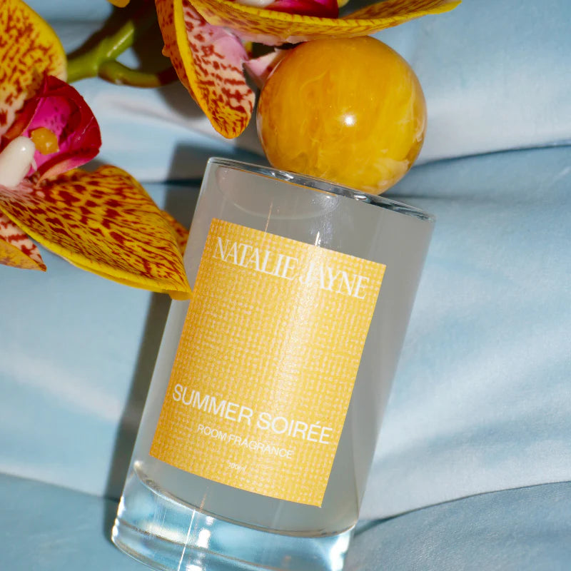 A clear glass perfume bottle labeled 'NATALIE LAINE SUMMER SOIRÉE' with a yellow-orange patterned label, styled with yellow orchid flowers and a yellow marble sphere on a light blue fabric background.
