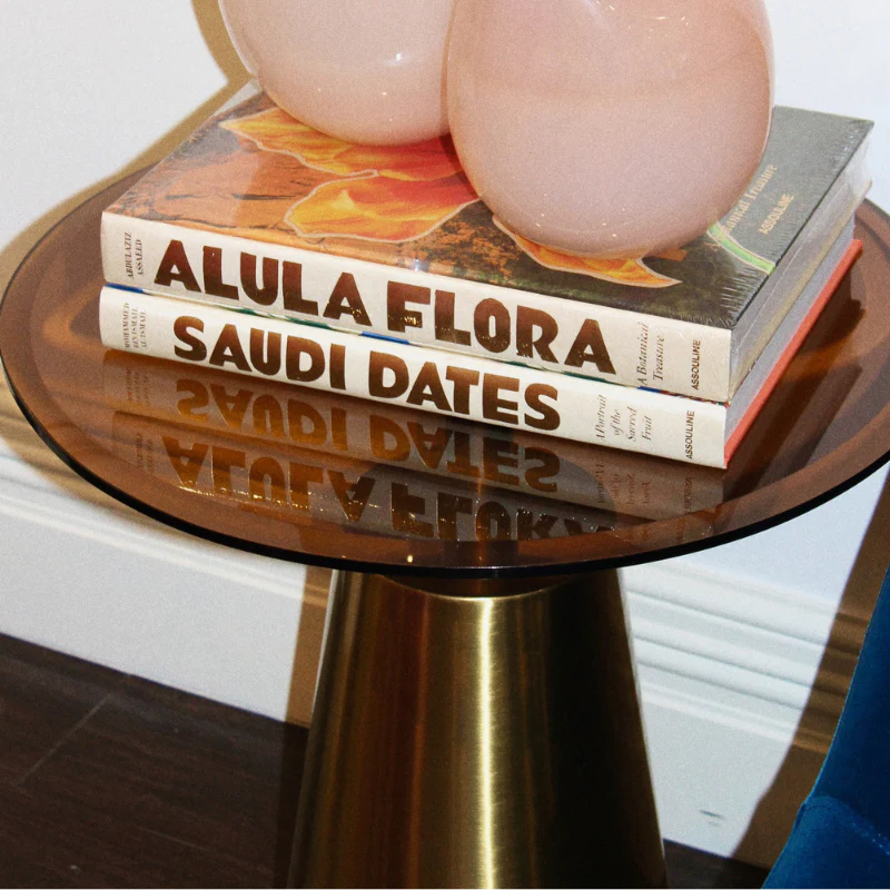 A round amber glass side table with a brushed brass pedestal base, styled with two pink spherical decor objects and stacked books including 'Alula Flora' and 'Saudi Dates'.