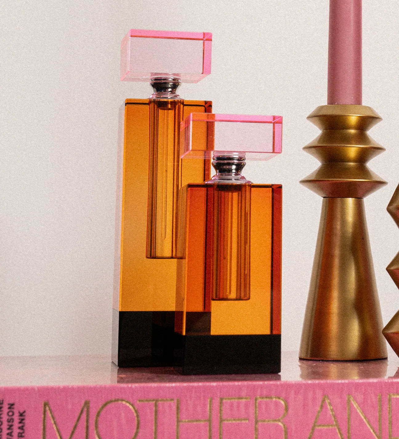 Two modern perfume bottles with translucent amber-orange bodies, black bases, and pink rectangular caps, styled on a pink book next to a gold candle holder.