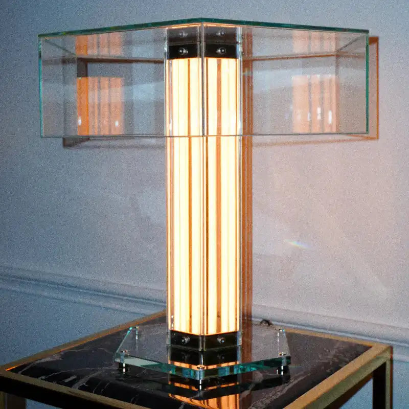 A modern table lamp with a glowing vertical glass column, a clear glass cross-shaped shade, and a square glass base, displayed on a dark marble-top table against a light gray wall.