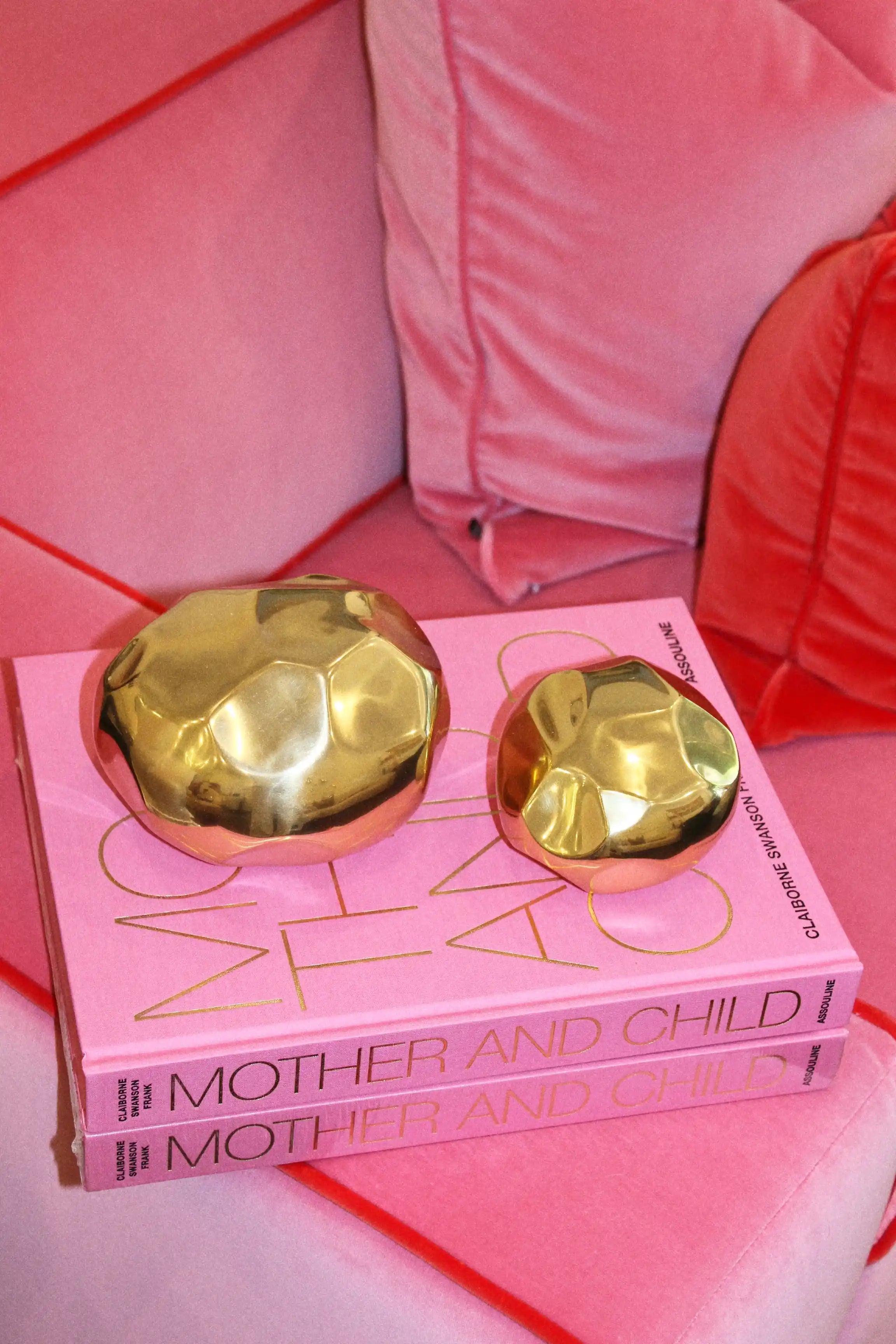 Two metallic gold decorative spheres resting on pink books titled 'MOTHER AND CHILD' on a pink sofa with pink cushions.