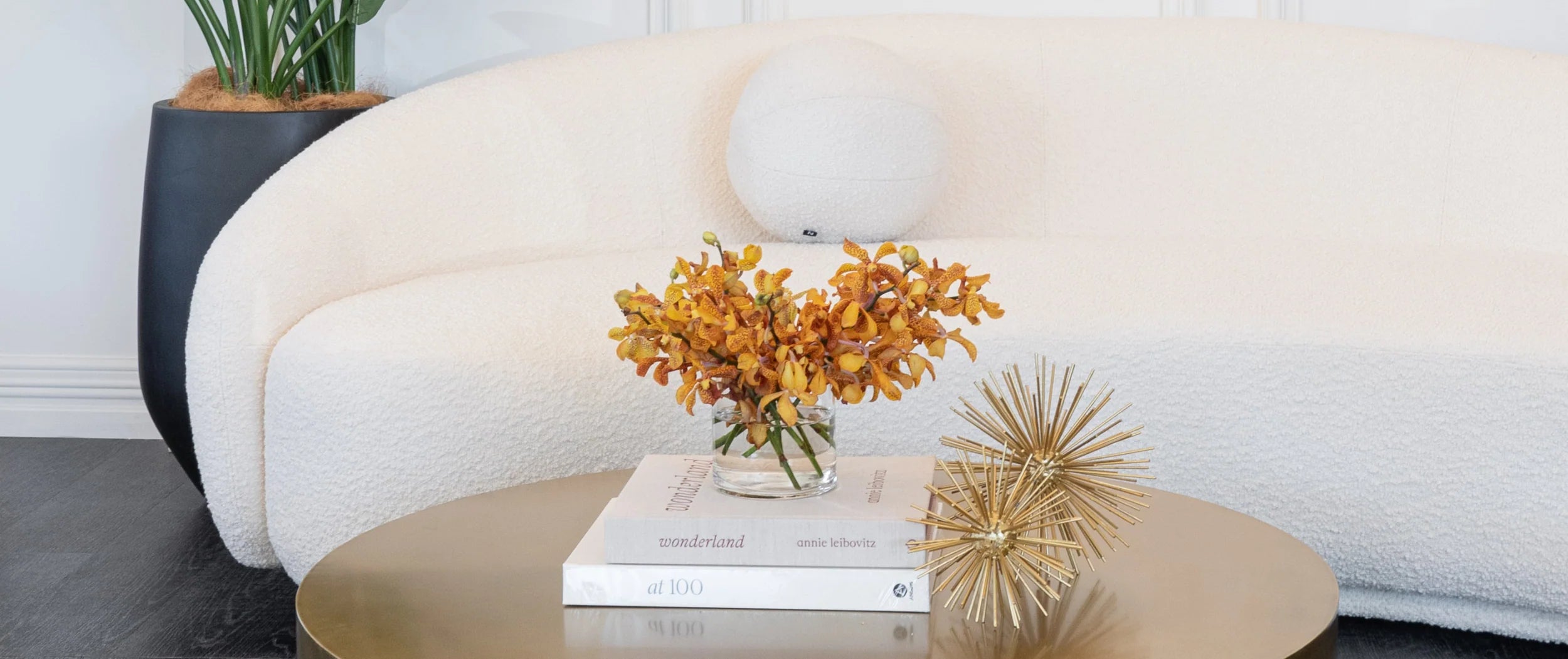 A styled interior scene showing a cream boucle sofa, a round gold coffee table, books, a vase of yellow-orange flowers, and a decorative gold spire, with a black planter in the background.