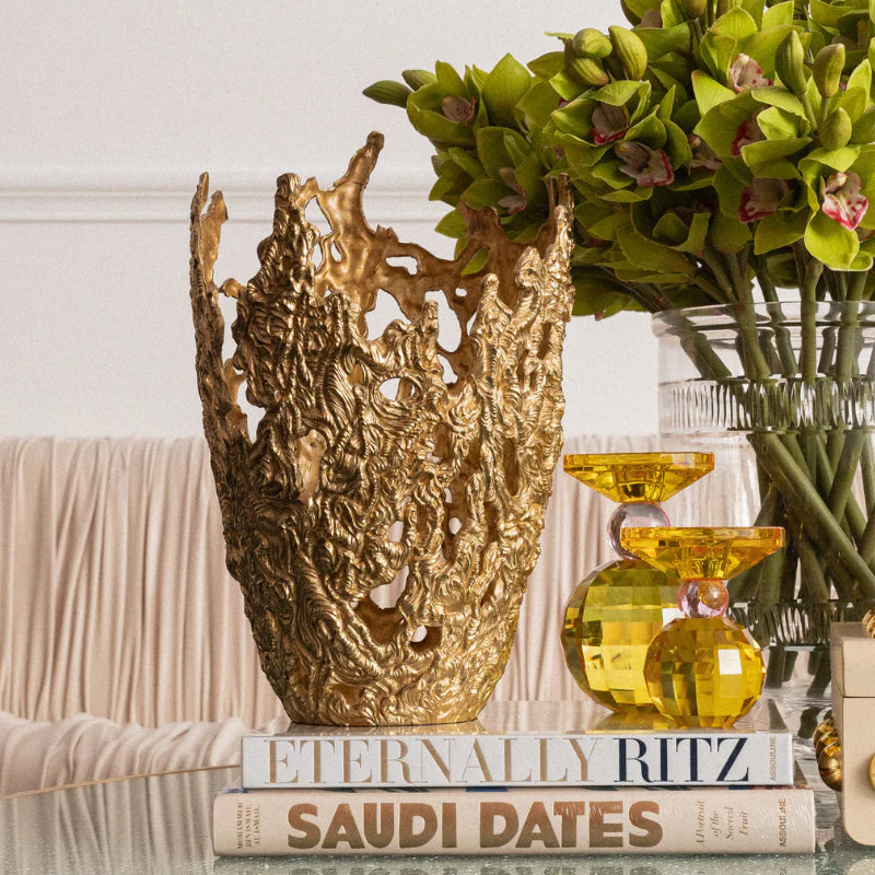 An ornate gold decorative vase with a highly textured, organic, coral-like silhouette, displayed on a stack of books next to two yellow glass hourglasses and a green orchid arrangement.