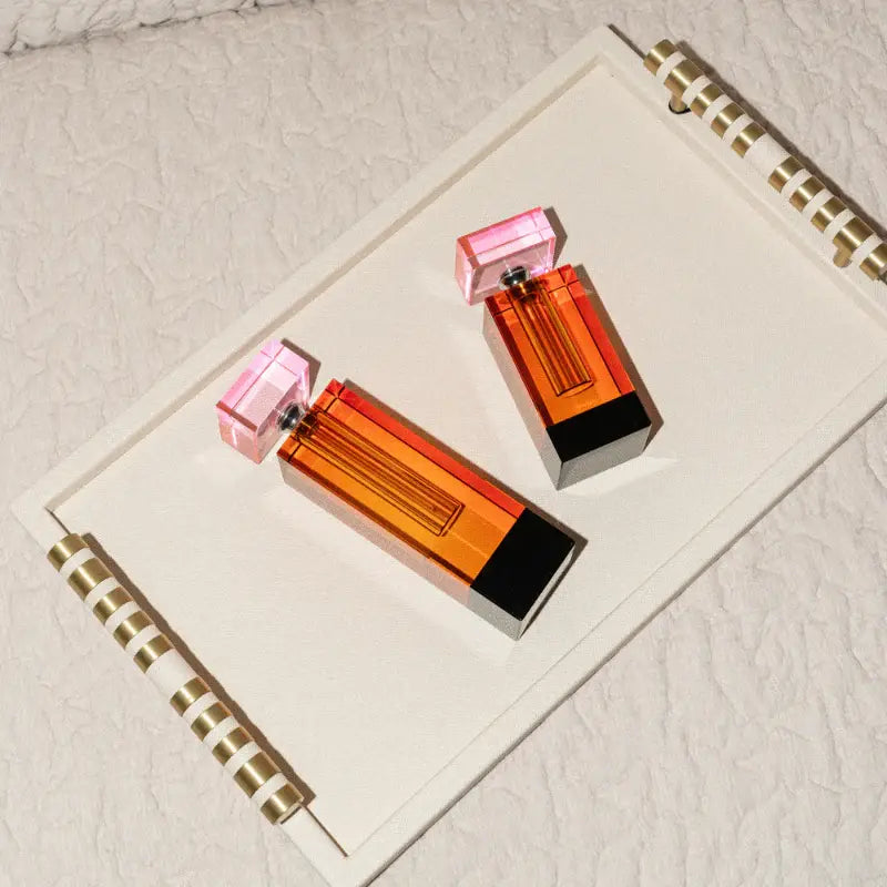 Two rectangular perfume bottles with orange and red gradient transparent bodies, pink faceted caps, and black bases, arranged on a white spiral-bound notebook against a textured off-white background.