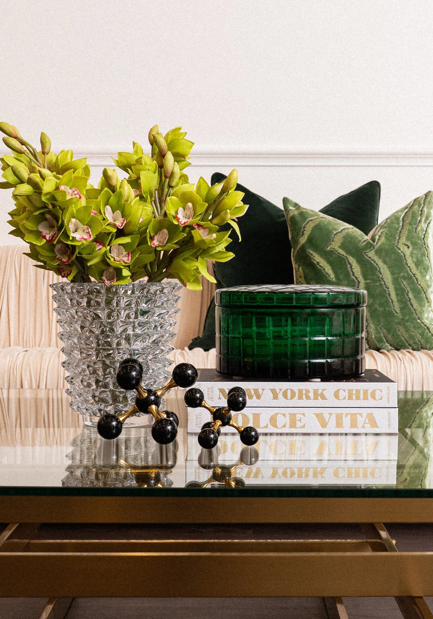 A styled interior scene showing a glass vase with bright green orchids, a textured green decorative box, stacked books, and a black-and-gold decorative object on a glass table in front of a sofa with green pillows.